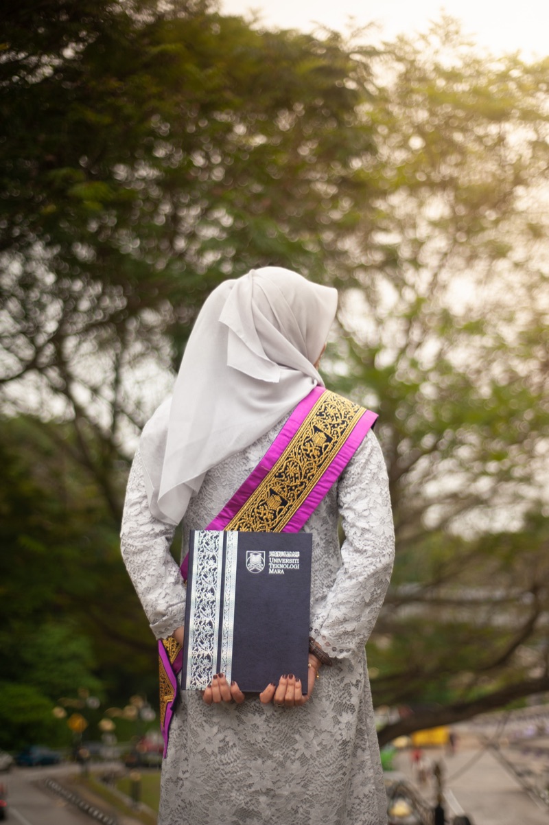 A graduate seen from behind, holding her certificate folder at sunset